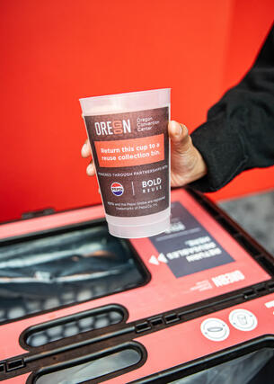 a heavy-duty plastic cup with the OCC logo and reuse messaging is being held directly over a red and black collection bin where guests place reusable cups and food baskets.