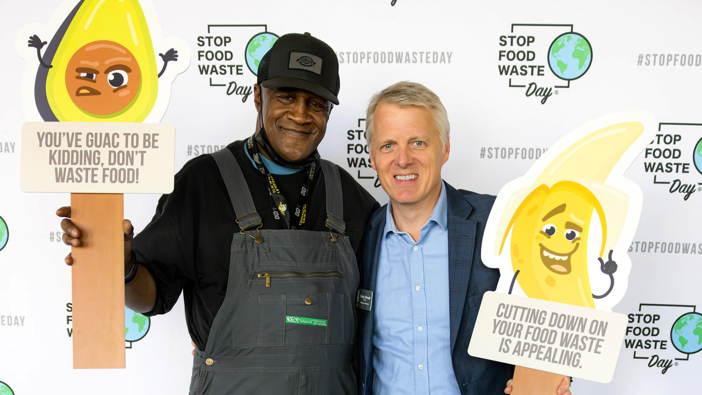 A man in overalls and a man in a blue suit stand next to each other holding food signs supporting "Stop Food Waste Day" 