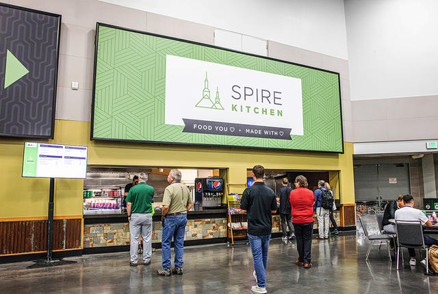 People near a food service counter filled with products. A large green sign hangs overhead.